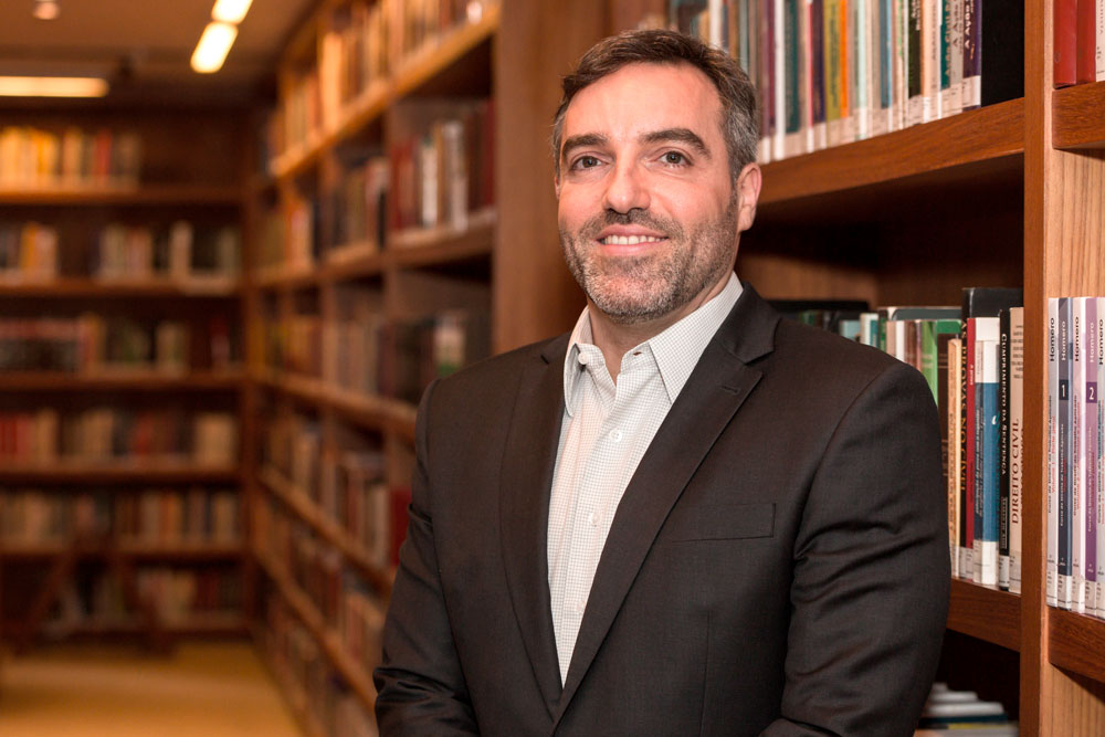 Retrato de Fábio Ozi em uma biblioteca. Ele está de pé, sorrindo, vestindo camisa branca e terno cinza-escuro. Ao fundo, há estantes com livros.