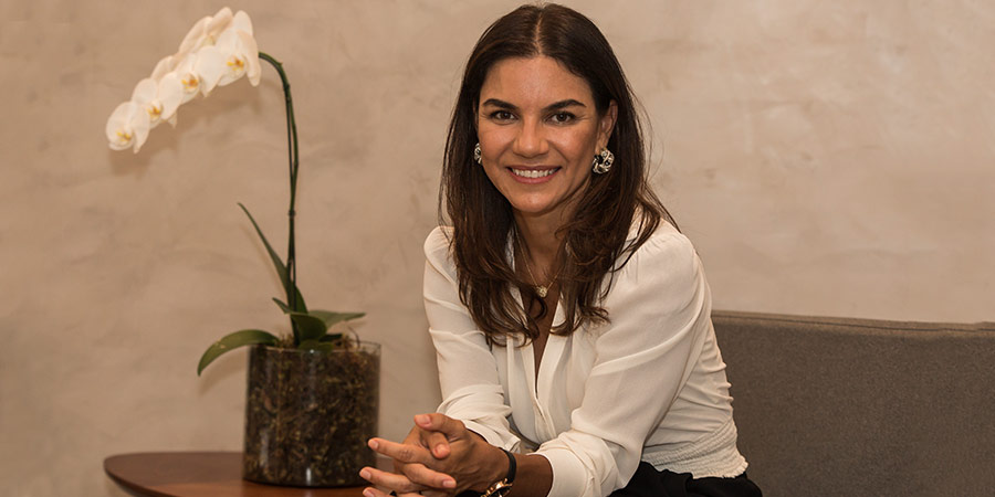 Retrato de Paula Vieira, sentada e sorrindo. Ela veste blusa branca, está levemente inclinada para frente, com os cotovelos apoiados nas coxas e as mãos entrelaçadas. Ao seu lado direito, vê-se uma mesa com um jarro de orquídeas.