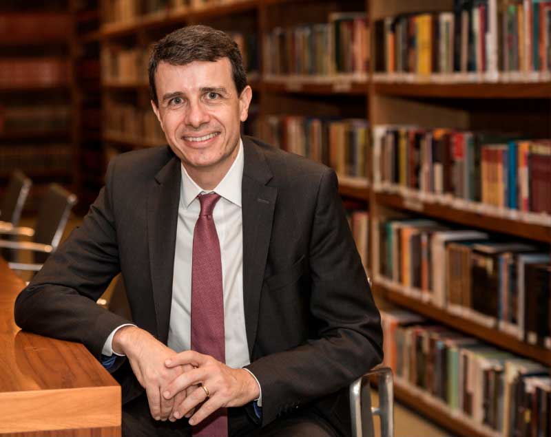 Retrato de Paulo Brancher em uma biblioteca. Ele está sentado com o braço direito apoiado em uma mesa e veste terno terno cinza-escuro, camisa cinza-claro e gravata vinho. Ao fundo, há estantes com livros.
