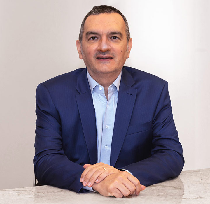 Retrato de Roberto Quiroga sentado, olhando para a frente e com um leve sorriso no rosto. Ele tem os antebraços apoiados sobre uma mesa branca, com uma mão sobre a outra. Ao fundo, há uma parede branca, e ele veste camisa social azul-clara e paletó também azul, de tom mais escuro.