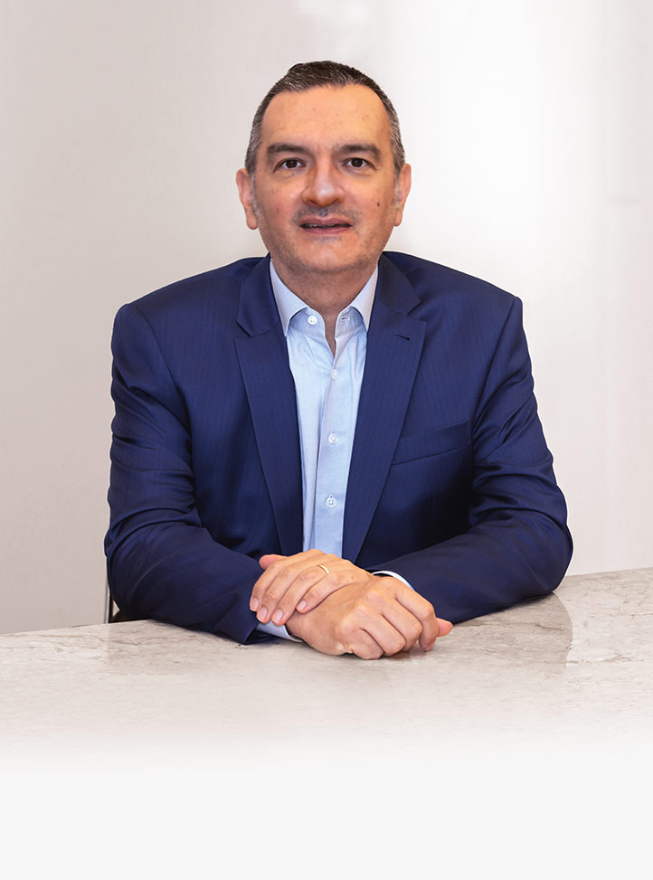 Retrato de Roberto Quiroga sentado, olhando para a frente e com um leve sorriso no rosto. Ele tem os antebraços apoiados sobre uma mesa branca, com uma mão sobre a outra. Ao fundo, há uma parede branca, e ele veste camisa social azul-clara e paletó também azul, de tom mais escuro.