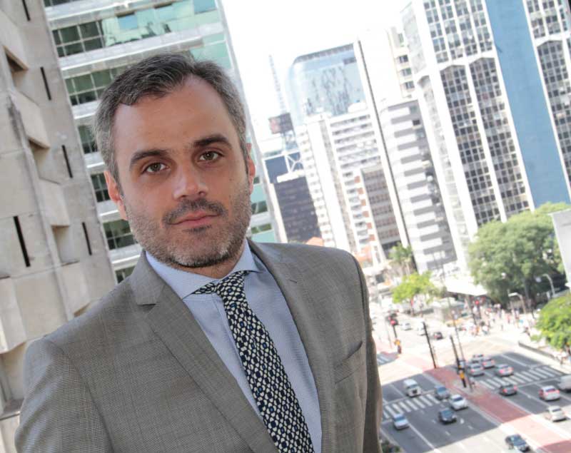Retrato de Rogério Taffarello, de pé, em uma sacada. Ele veste terno cinza-claro, camisa branca e gravata com estampa geométrica em preto e branco. Ao fundo, vê-se a Avenida Paulista.