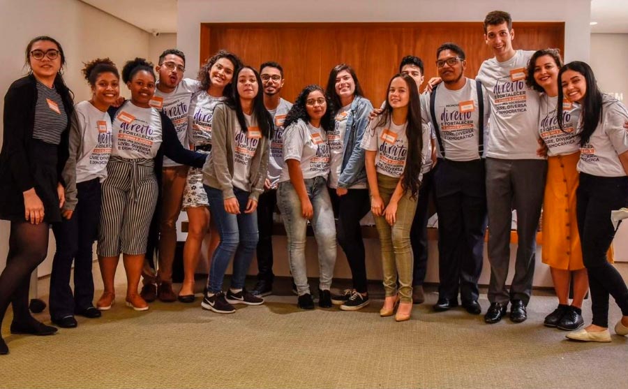 Foto de um grupo de 15 jovens, sendo 10 mulheres e cinco homens. Eles posam lado a lado, sorrindo e vestindo camisetas do programa de bolsas.